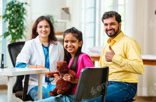 Preview: Happy little Indian girl holding teddy bear while sitting in hospital as father, doctor observe