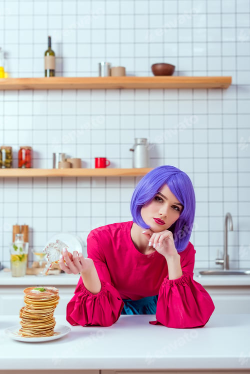 Preview: housewife with purple hair and colorful clothes pouring syrup on pancakes while looking at camera in