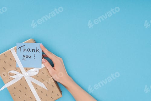 Preview: cropped view of hand and sticky note with thank you lettering on gift box isolated on blue