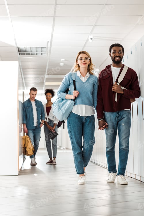 Preview: full length view of smiling multiethnic students holding hands in college