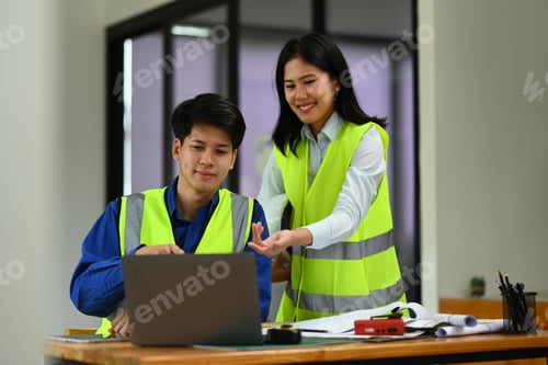 Preview: Two engineers in reflective vests working on blueprint and discussing the new project in office