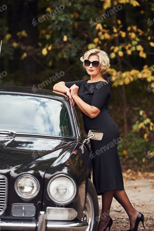 Preview: Blonde woman in sunglasses and in black dress near old vintage classic car