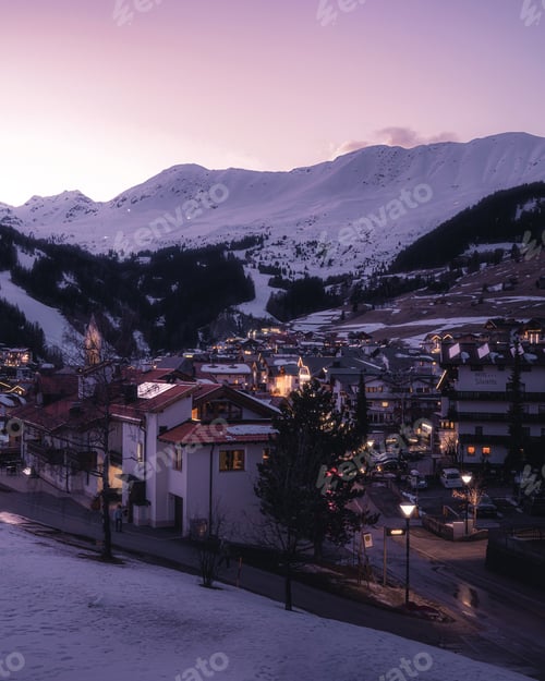 Preview: Vertical shot of dense buildings with illuminated lights and mountains under a clear sky in winter
