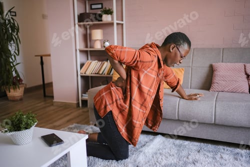 Preview: Young woman suffering back pain at home, kneeling on the floor