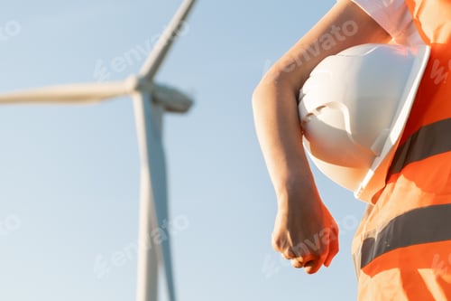 Preview: An engineer with a helmet in his hands and orange vesta stands on the background of windmill or wind