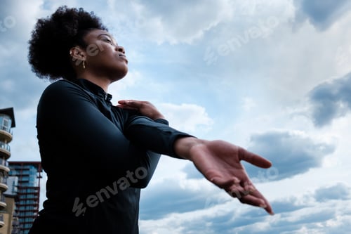 Preview: Portrait of fit young black woman stretching with a cloudy sky on the background.