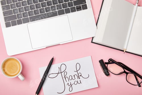 Preview: top view of laptop, coffee, notebook, glasses and card with thank you lettering at workplace on pink