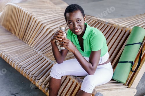Preview: African woman with short hair smiling holding reusable bottle sitting on bench with yoga mat