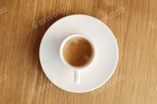 Vorschau: Ansicht von oben auf eine weiße Porzellantasse mit einem Teller mit heißem Kaffee mit Milch auf einem Holztischhintergrund.