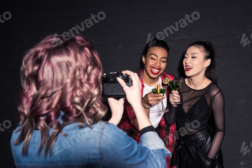 Preview: friend taking photo of smiling multiethnic friends with cocktails at party