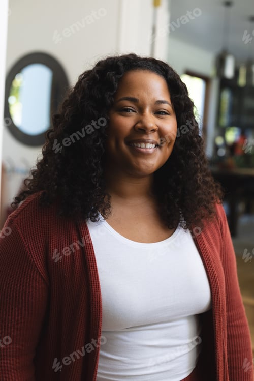 Preview: Happy african american woman with curly hair standing and smiling in sunny house