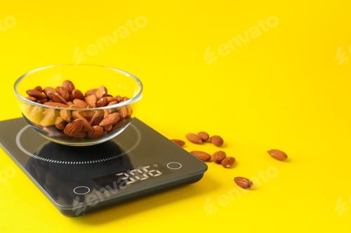 Preview: Electronic kitchen scale with bowl of almond nuts on yellow background, closeup. Space for text