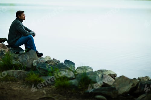 Preview: A middle-aged man sitting on rocks looking pensively over the water.