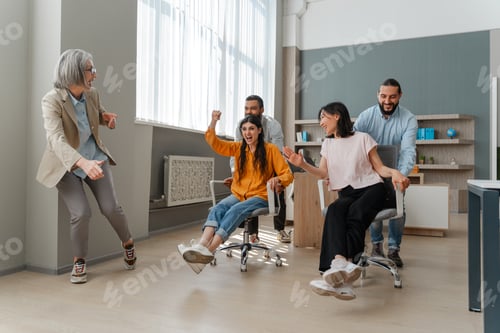 Preview: Happy business team having fun racing on chairs in office