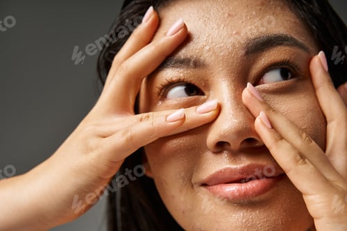 Preview: happy young asian woman with brunette hair and blemishes on wet face on grey background, close up