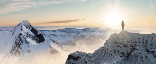 Preview: Adventurous Woman Hiker standing on top of icy peak with rocky mountains in background.