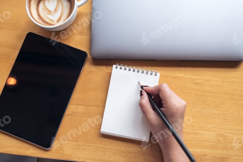 Preview: a woman's hand holding pencil to writing on blank notebook with tablet , laptop and coffee