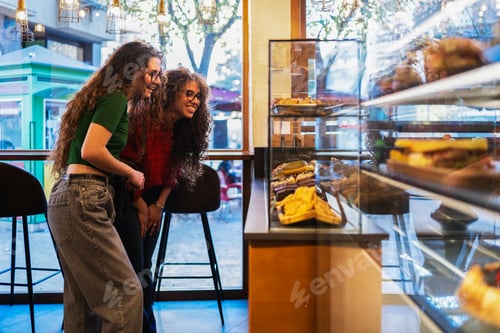 Preview: Two smiling friends choosing pastries in a modern cafe