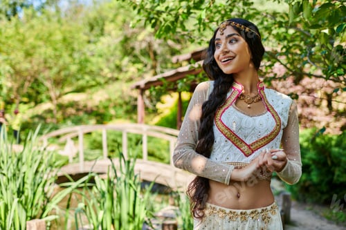 Preview: amazed and happy indian woman in elegant ethnic wear looking away in green summer park