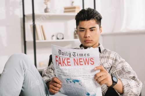 Preview: serious asian man sitting in sofa at home and reading fake news newspaper