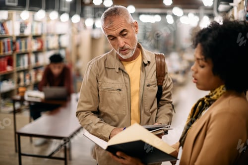Preview: Mature student and his black female professor examining reading book in a library.