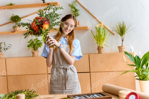 Preview: beautiful young florist in eyeglasses holding flower bouquet and smiling at camera at workplace