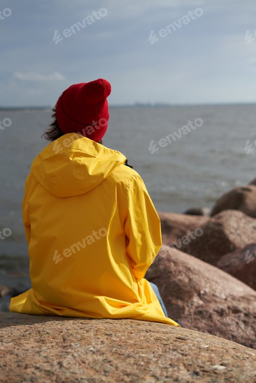 Preview: Back view of woman sitting in bright yellow cloak and red hat on stone near shore of North sea