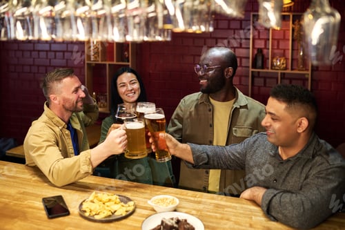 Preview: Cheerful intercultural friends with glasses of beer clinking over bar counter