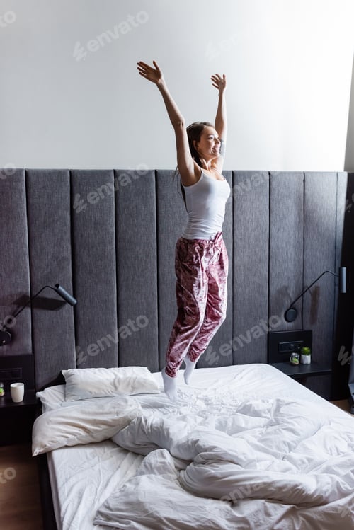 Preview: cheerful young woman with hands above head jumping on bed