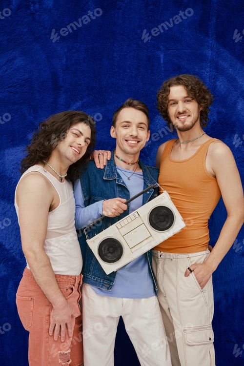 Preview: three modish joyous gay friends posing with tape recorder on dark blue backdrop, pride month