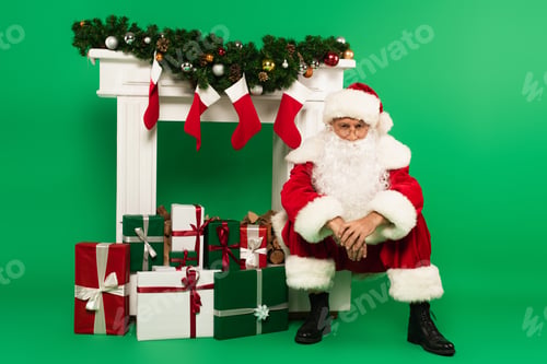 Preview: Santa claus looking at camera near gifts and decorated fireplace on green background