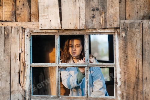 Preview: beautiful girl looks out the window from an old wooden house, evening sunlight
