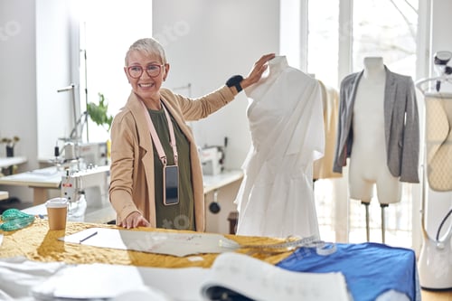 Preview: Smiling mature fashion designer stands between mannequin and cutting table in sewing workshop