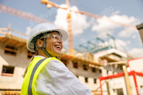Preview: Smiling female construction worker in protective helmet standing against on construction background