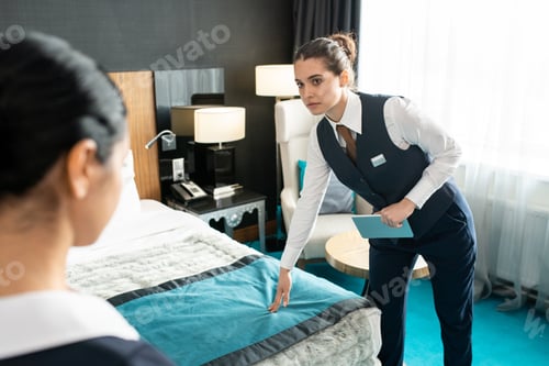 Preview: Young serious hotel worker holding digital tablet while touching bed and looking at new room maid