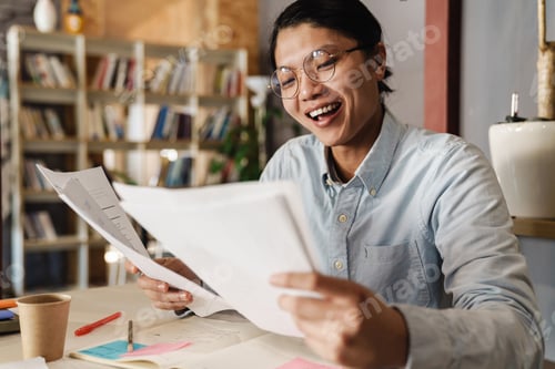 Preview: Image of happy asian man in eyeglasses laughing while doing homework