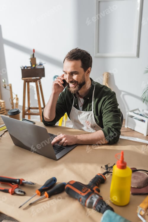 Preview: Cheerful carpenter talking on smartphone and using laptop near tools in workshop