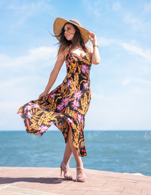 Preview: Vertical shot of a female in a floral sundress and a hat standing by the sea captured in Spain