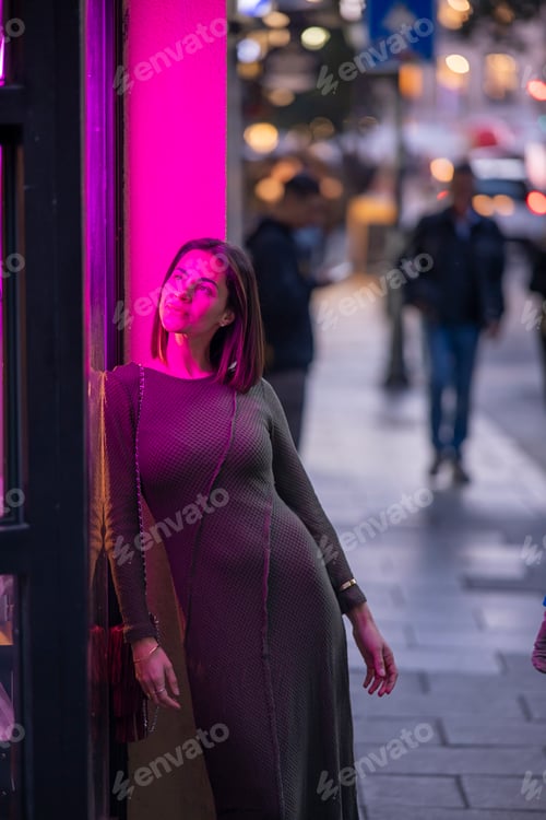 Preview: Urban portrait of woman illuminated by vibrant magenta streetlights, standing near glass.