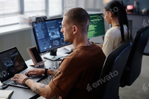 Preview: Side View of Male IT Programmer with Earring Coding on Laptop at Working Desk in Office