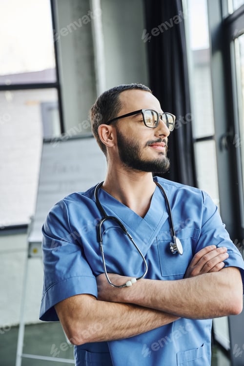 Preview: dreamy and smiling professional paramedic in eyeglasses and blue uniform standing with folded arms