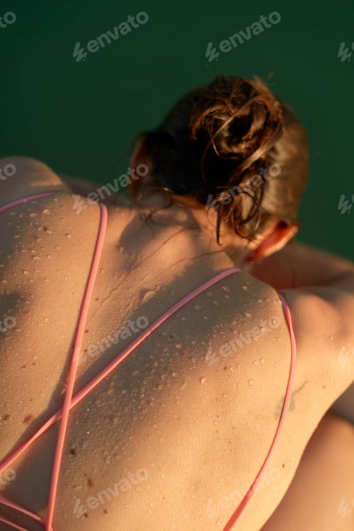 Preview: Sunlit back of a person with wet hair in a pink swimsuit sitting by the water. Brandenburg, Germany