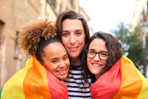 Preview: Three lesbian and trans women are hugging each other wrapped in a rainbow flag.