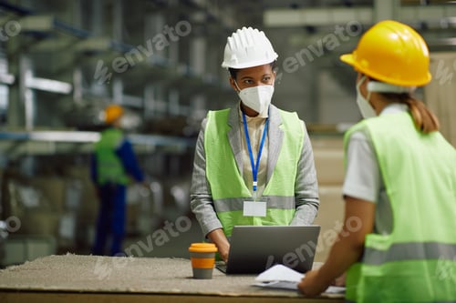 Preview: Black female supervisor working on laptop and talking to warehouse worker while wearing face mask.