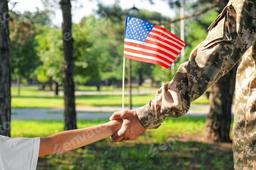 Preview: Soldier and his little son with American flag holding hands outdoors, closeup. Veterans Day in USA