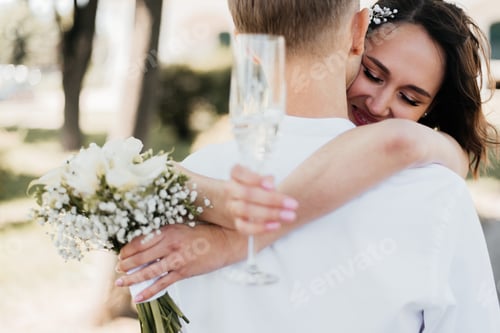 Preview: Cropped view of glass of champagne in hand of blurred bride and groom outdoors