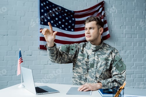 Preview: handsome man in military uniform pointing with finger and sitting near laptop