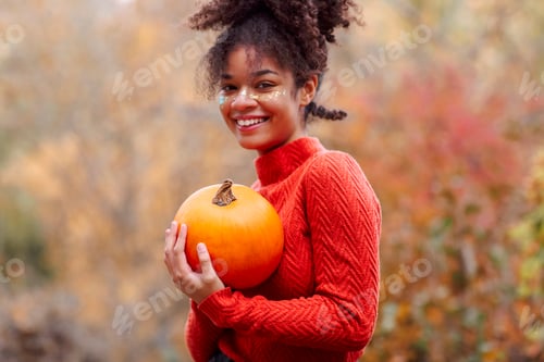 Preview: Young happy cheerful african american woman with curly hair with orange pumpkin in autumn forest