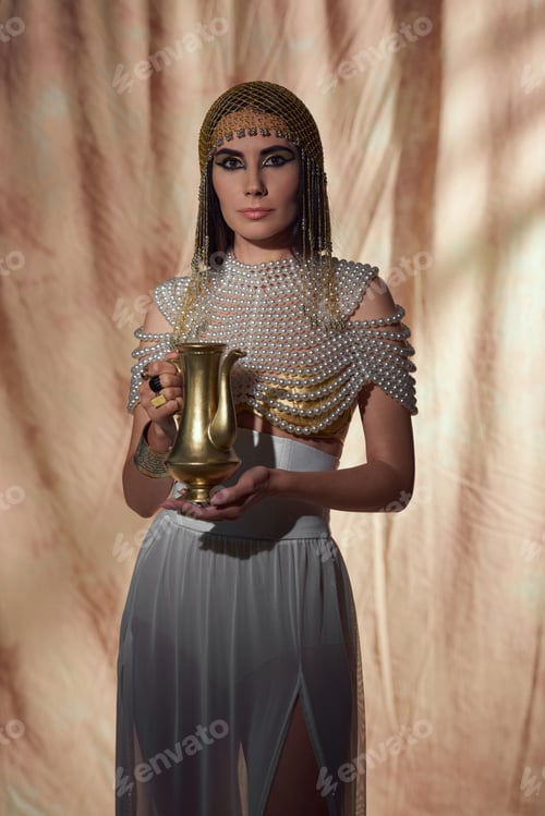 Preview: Elegant woman in egyptian look and pearl necklace holding shiny jug on abstract background