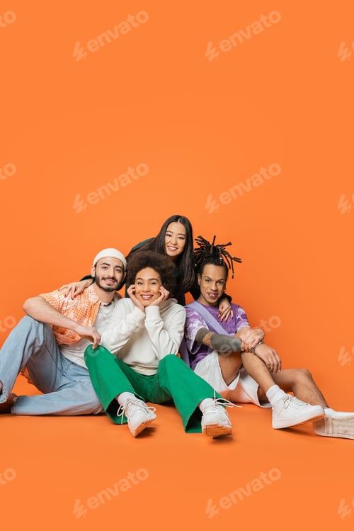 Preview: multiethnic friends in trendy clothes sitting and smiling at camera on orange background